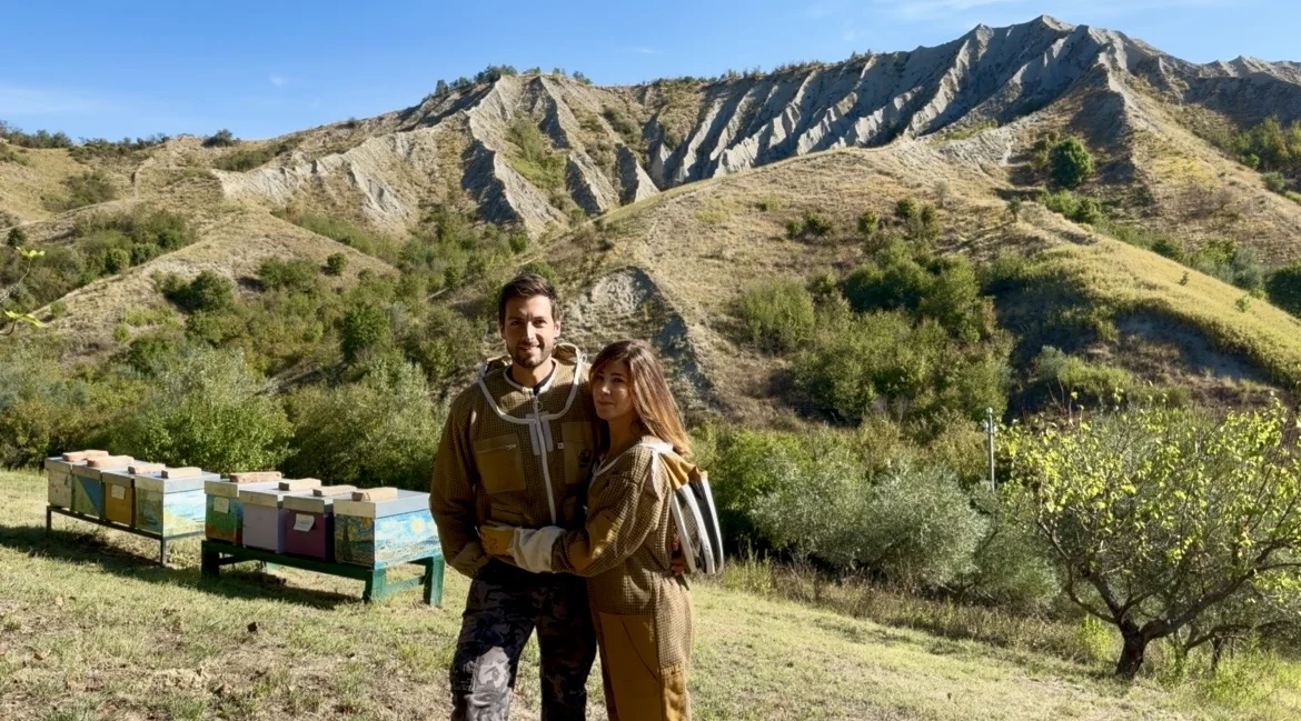 Lisa and Filippo smiling in their beekeeping suits, with the hives and the badlands behind them